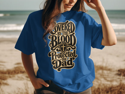 Blue t-shirt with text "Covered By The Blood Protected By Dad" and a shield with a cross.