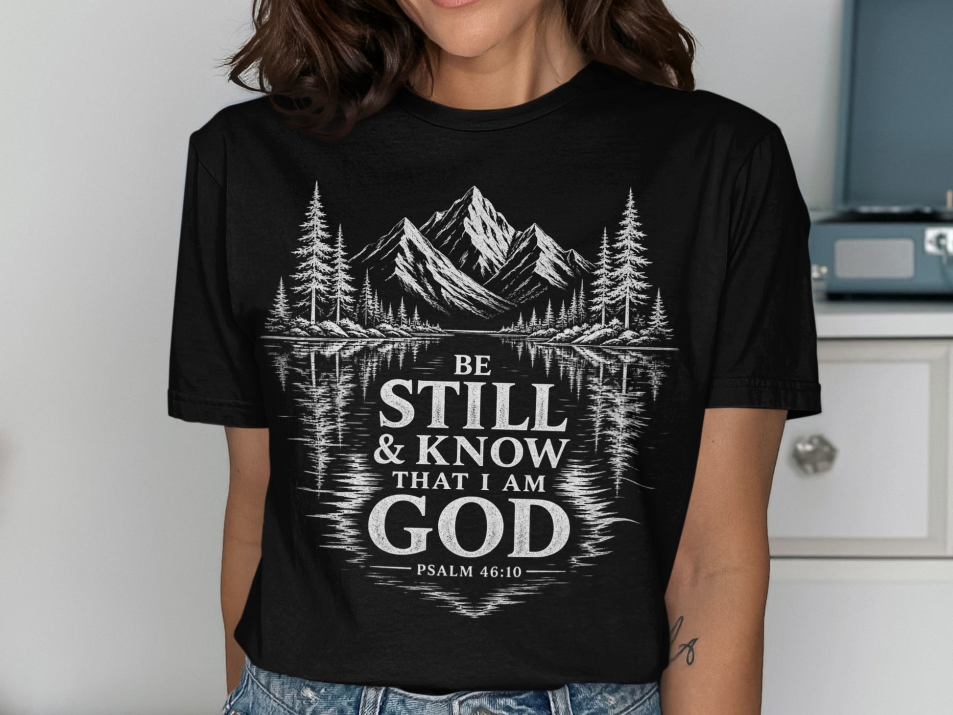 Black t-shirt with white graphic of mountains, trees, and text "BE STILL & KNOW THAT I AM GOD PSALM 46:10".