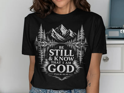 Black t-shirt with white graphic of mountains, trees, and text "BE STILL & KNOW THAT I AM GOD PSALM 46:10".