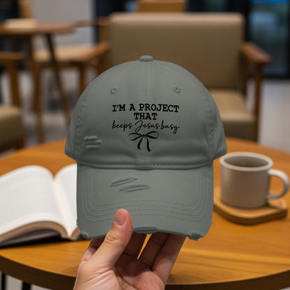 Distressed gray baseball cap with "I'm A Project That Keeps Jesus Busy" text and bow.
