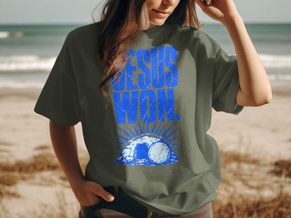 T-shirt with blue "Jesus Won" text over a tomb graphic on a beach.