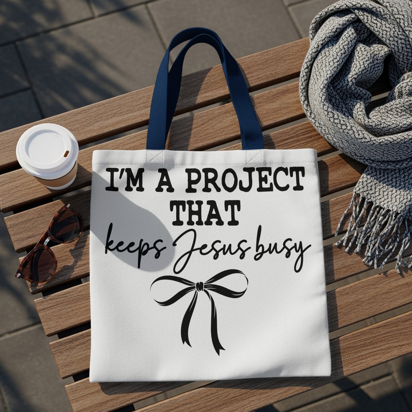 White tote bag with black text that reads "I'm a project that keeps Jesus busy."