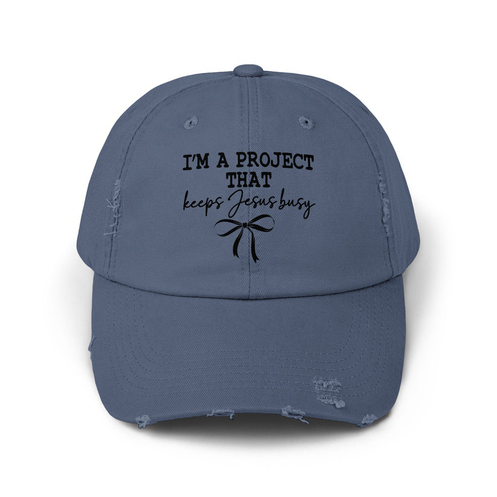 Distressed blue baseball cap with black text "I'M A PROJECT THAT keeps Jesus busy" and a bow.