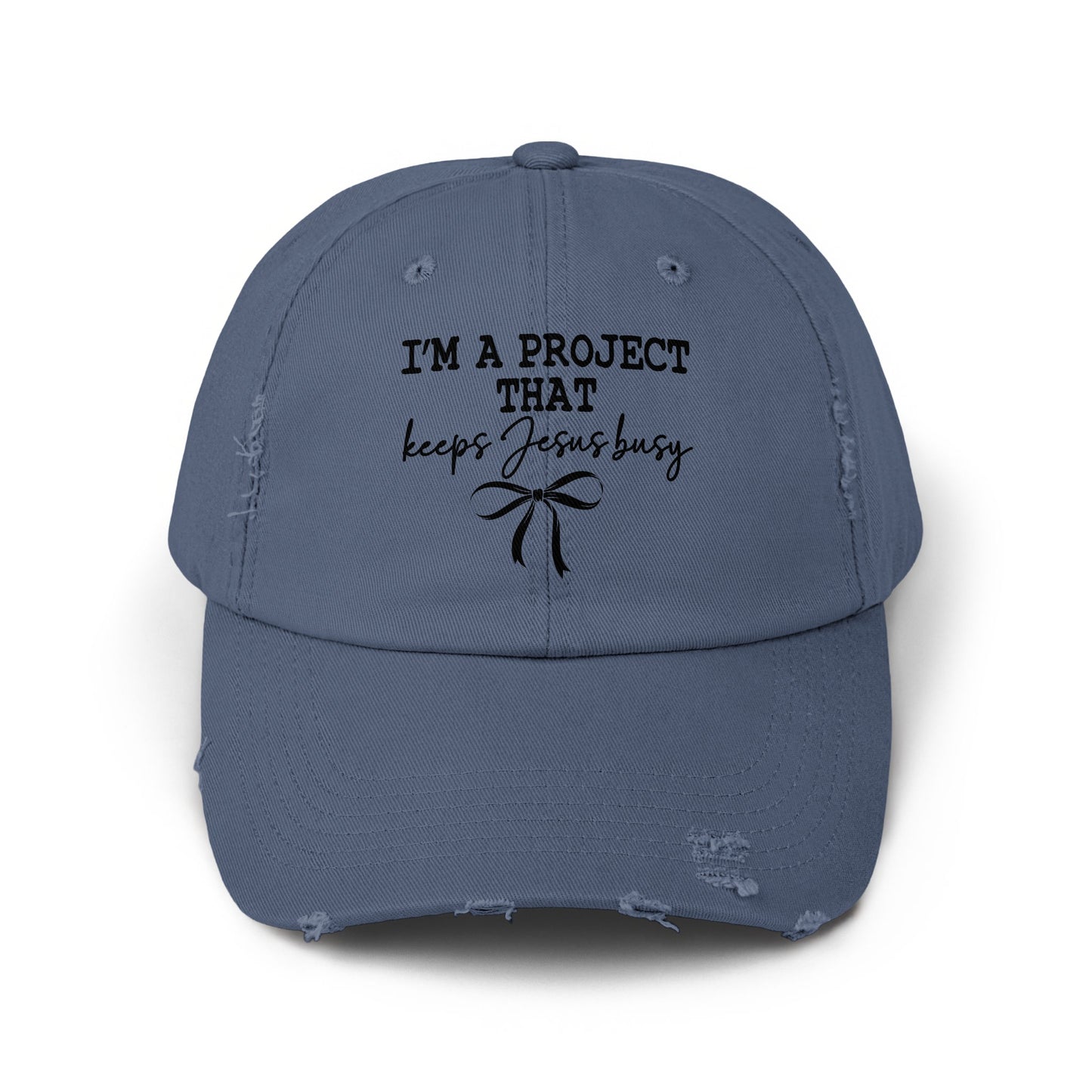 Distressed blue baseball cap with black text "I'M A PROJECT THAT keeps Jesus busy" and a bow.