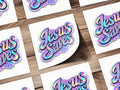 Holographic sticker with iridescent "Jesus Saves" text on a checkerboard background.