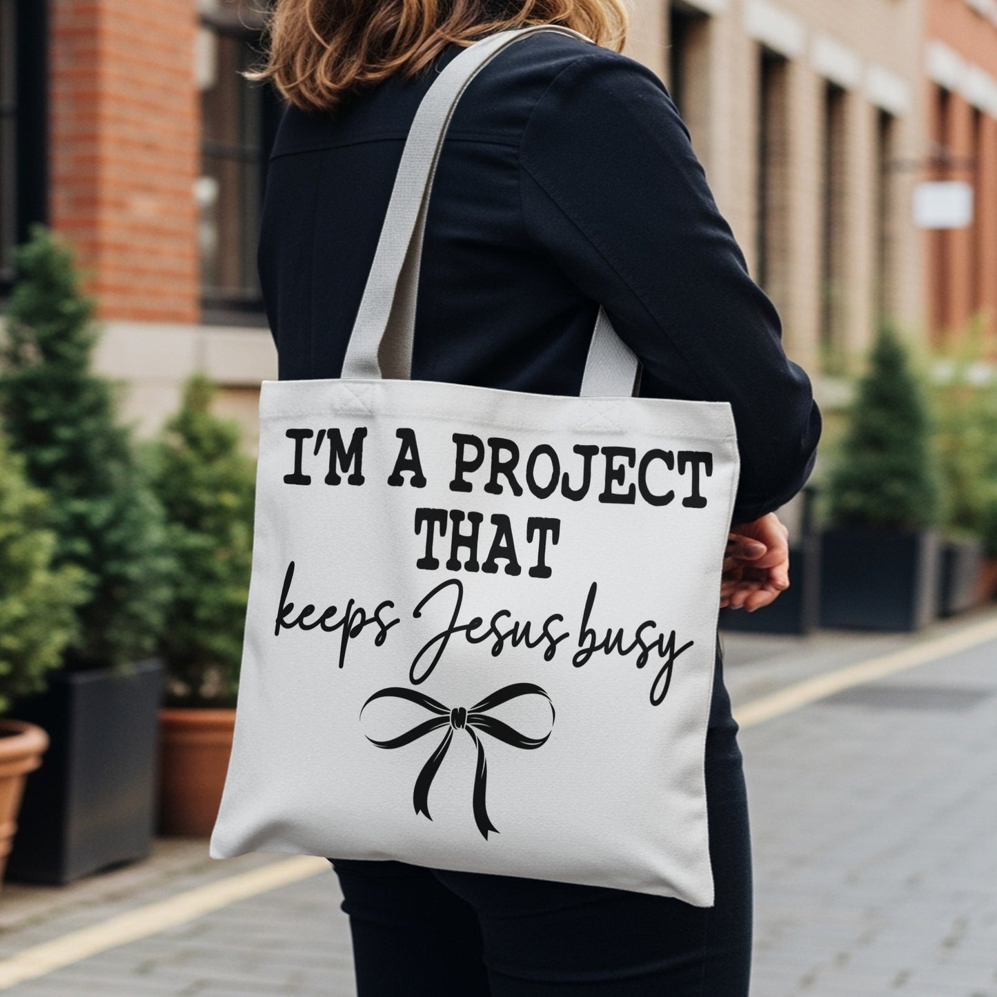 White tote bag with black text: "I'M A PROJECT THAT keeps Jesus busy", and a bow graphic.
