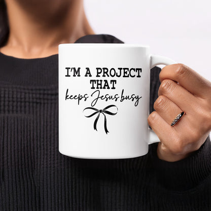 White mug with "I'm a project that keeps Jesus busy" and bow graphic.