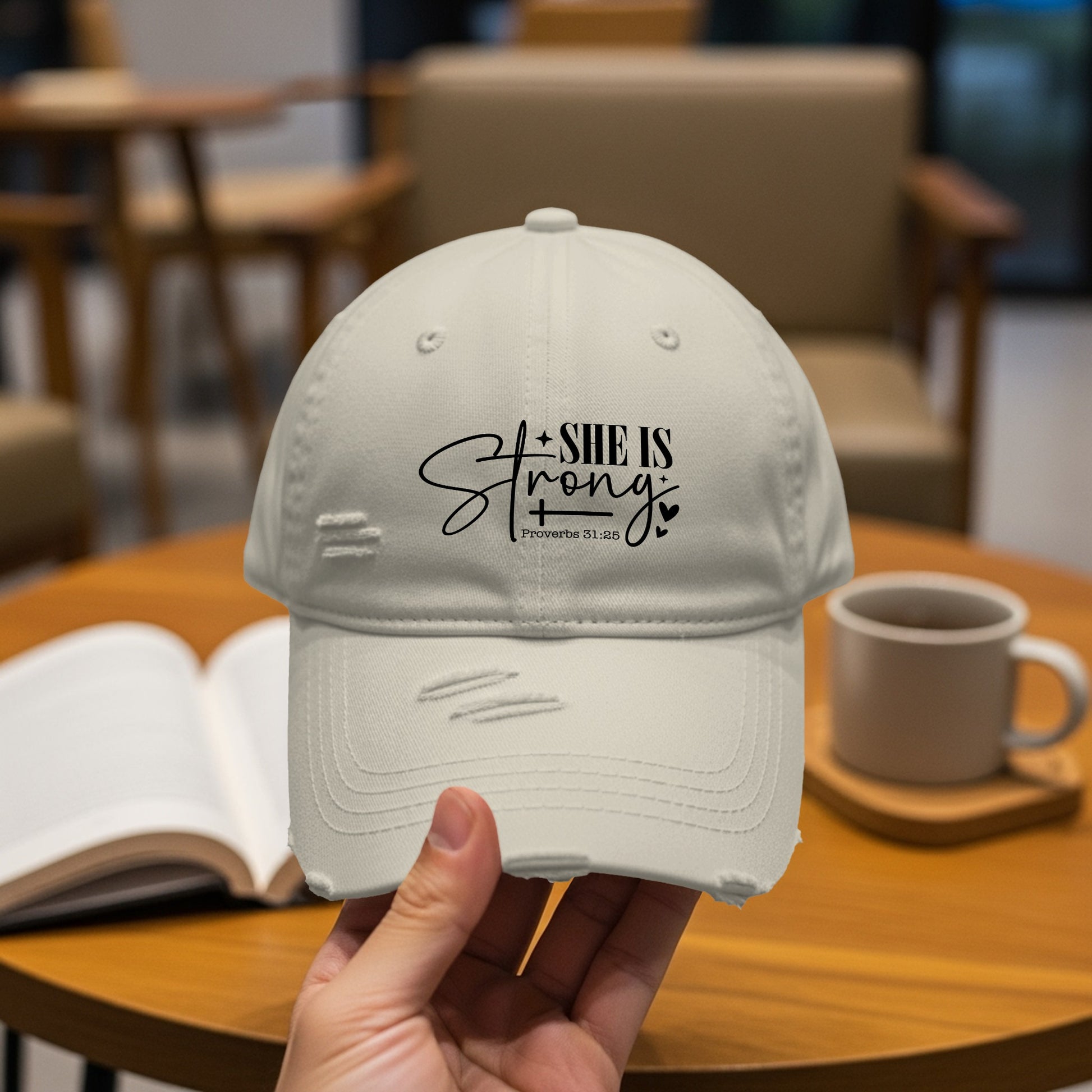 Light beige distressed baseball cap with black text "SHE IS Strong Proverbs 31:25".