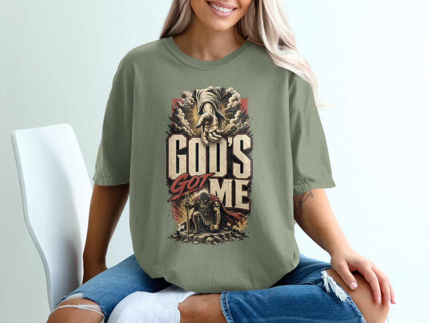 Olive green t-shirt with "God's Got Me" graphic featuring a hand reaching down and a kneeling soldier.