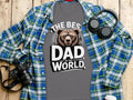 Grey t-shirt with "The Best Dad in the World" text and bear graphic, on a wooden surface with headphones and camera.
