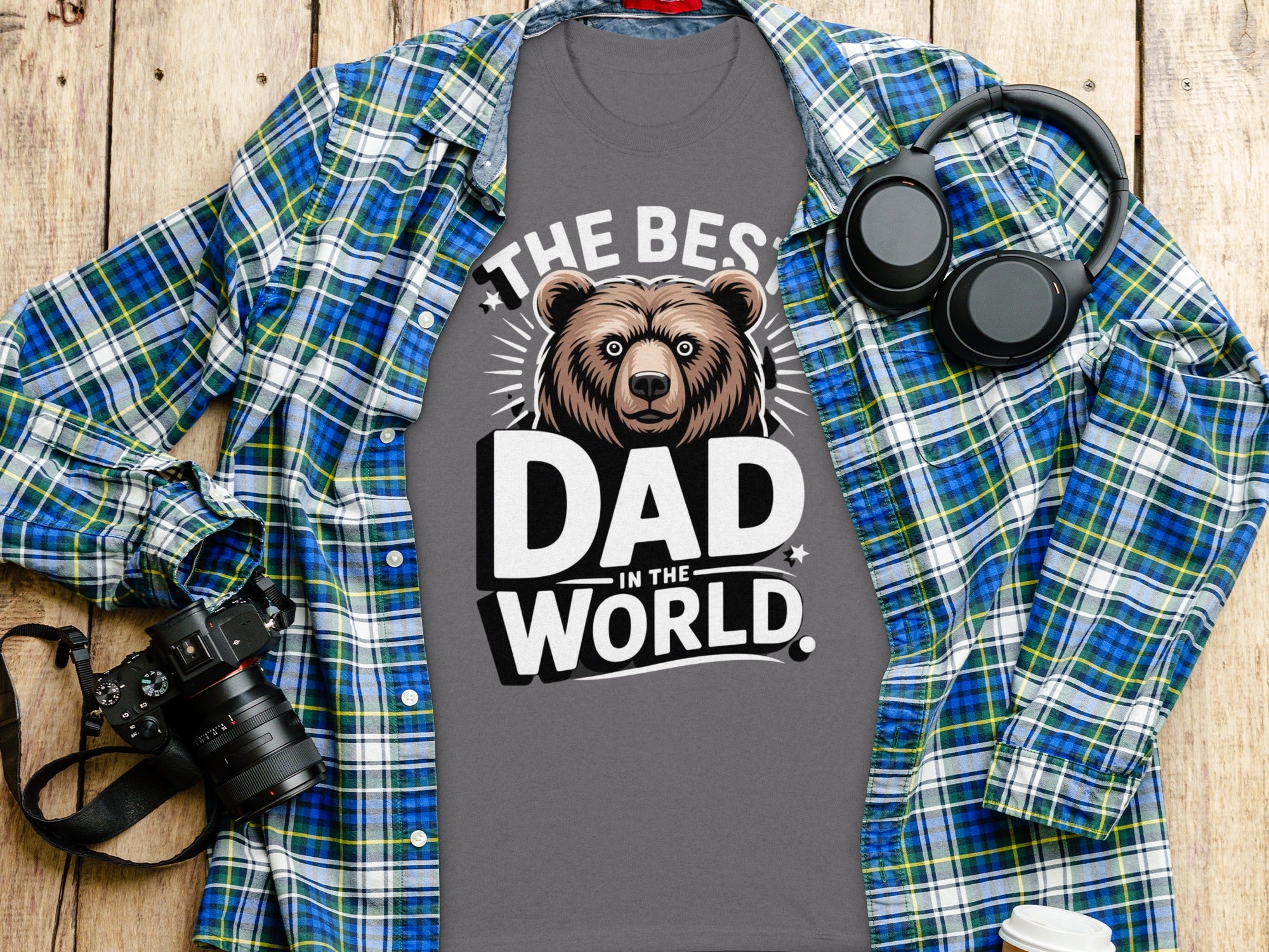 Grey t-shirt with "The Best Dad in the World" text and bear graphic, on a wooden surface with headphones and camera.