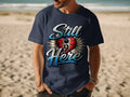 Navy t-shirt with "Still Here" text and a heart with a cross graphic.