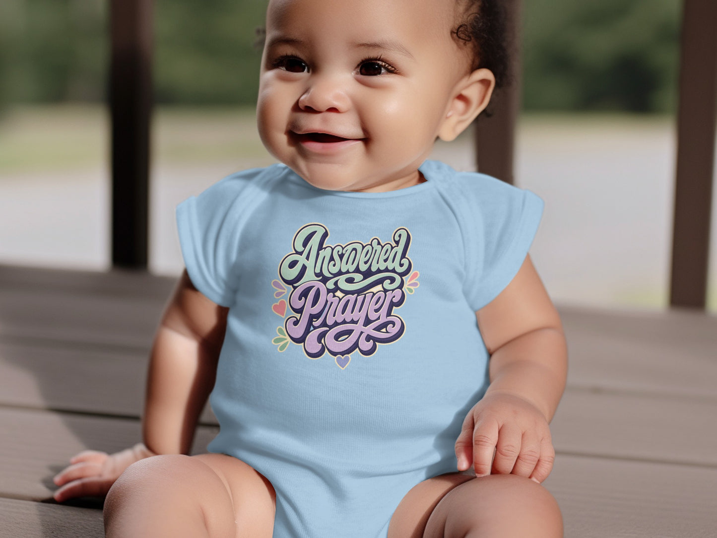 Baby in light blue onesie with "Answered Prayer" text and pastel hearts design.