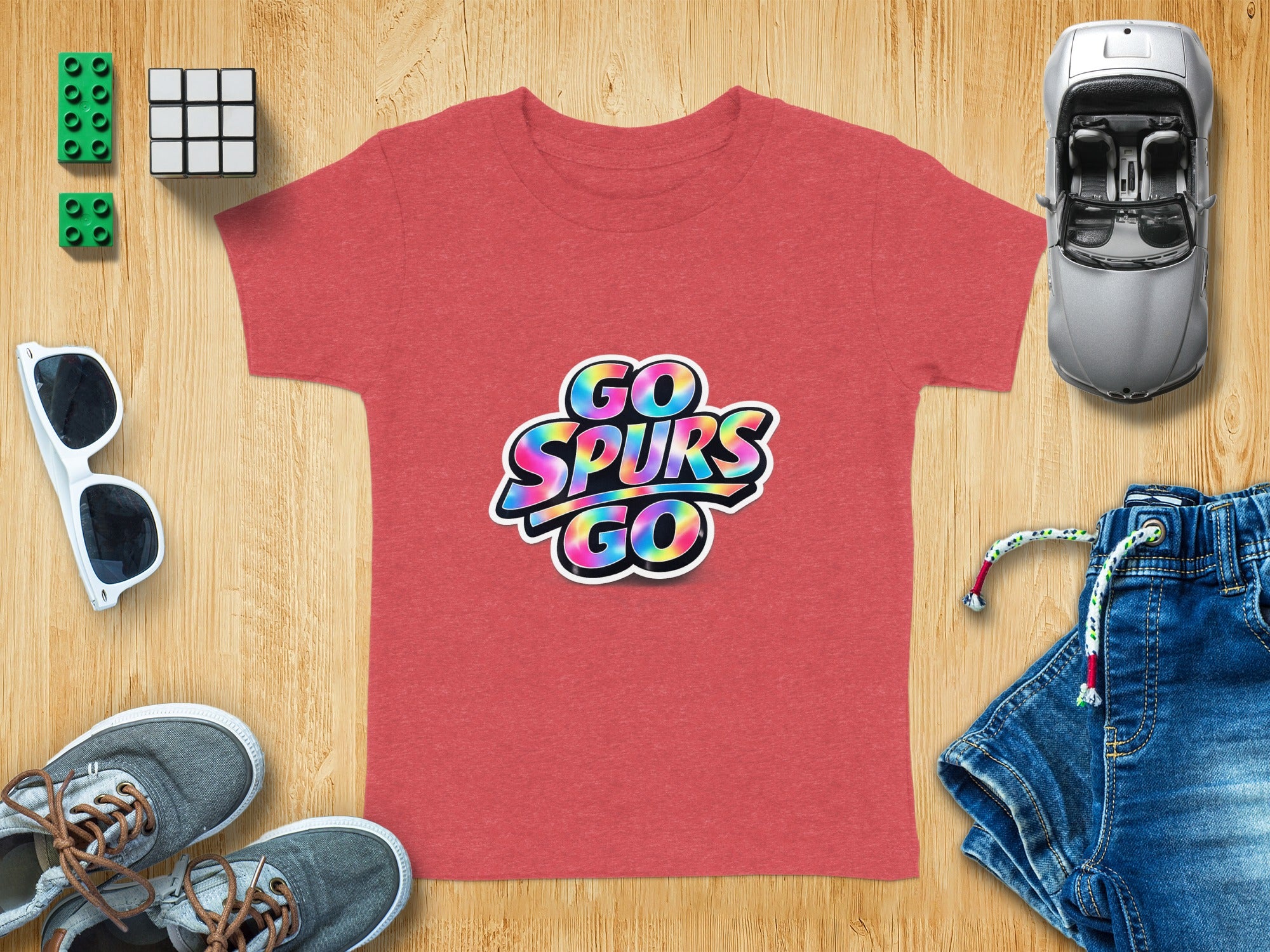 Red t-shirt with rainbow "GO SPURS GO" graphic and surrounding accessories on wood.