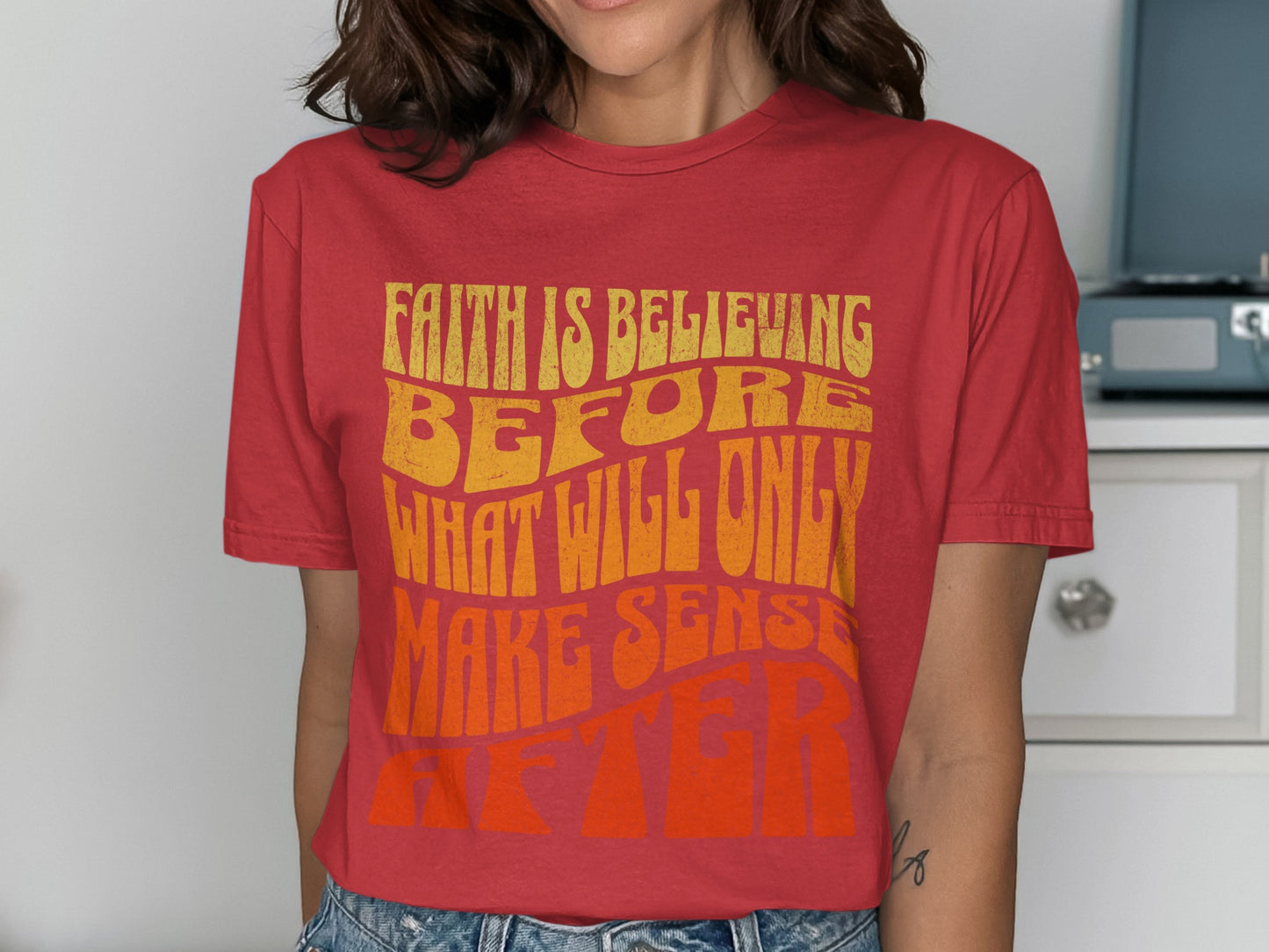 Red t-shirt with yellow and orange text reading "FAITH IS BELIEVING BEFORE WHAT WILL ONLY MAKE SENSE AFTER"