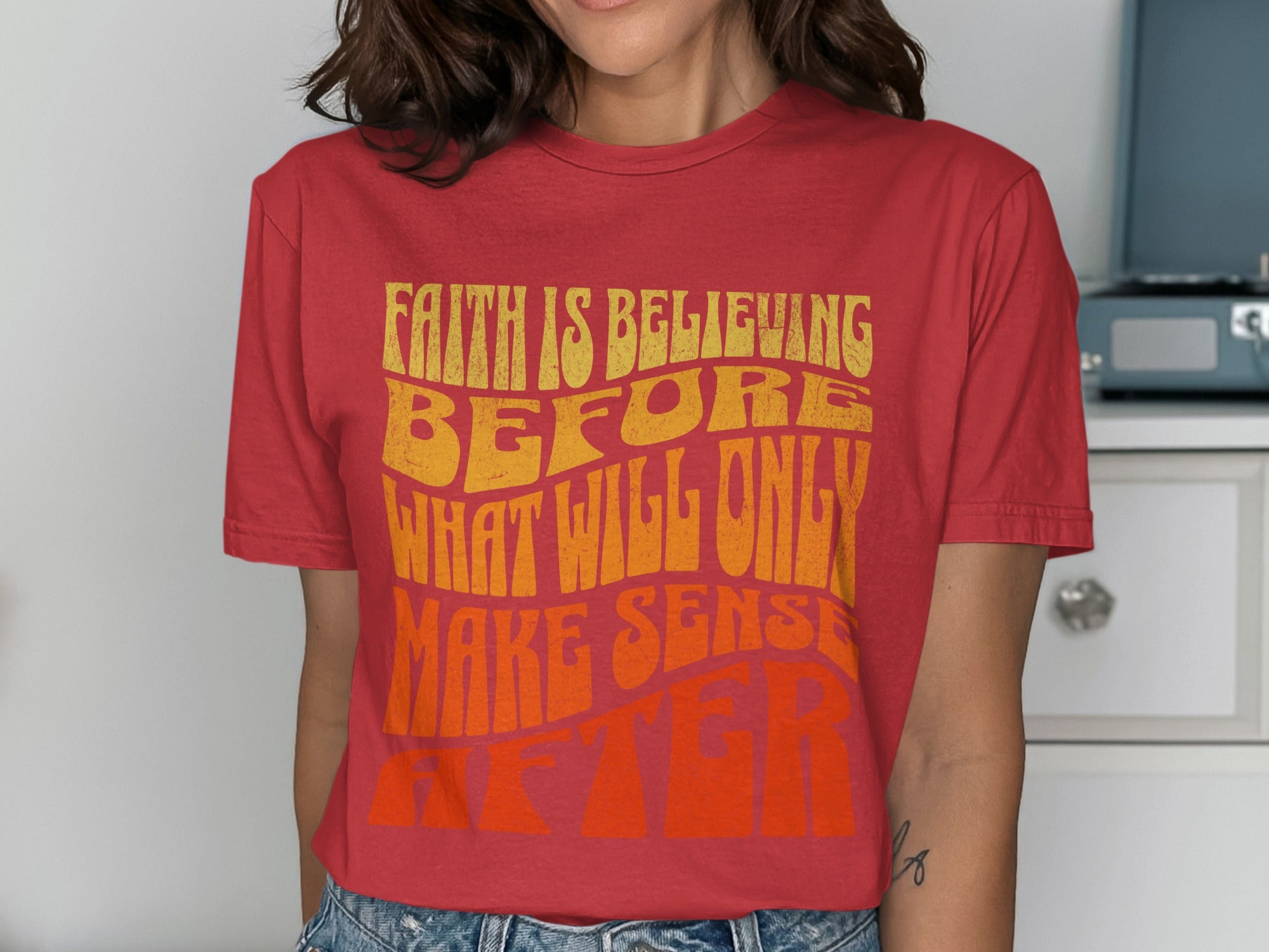 Red t-shirt with yellow and orange text reading "FAITH IS BELIEVING BEFORE WHAT WILL ONLY MAKE SENSE AFTER"