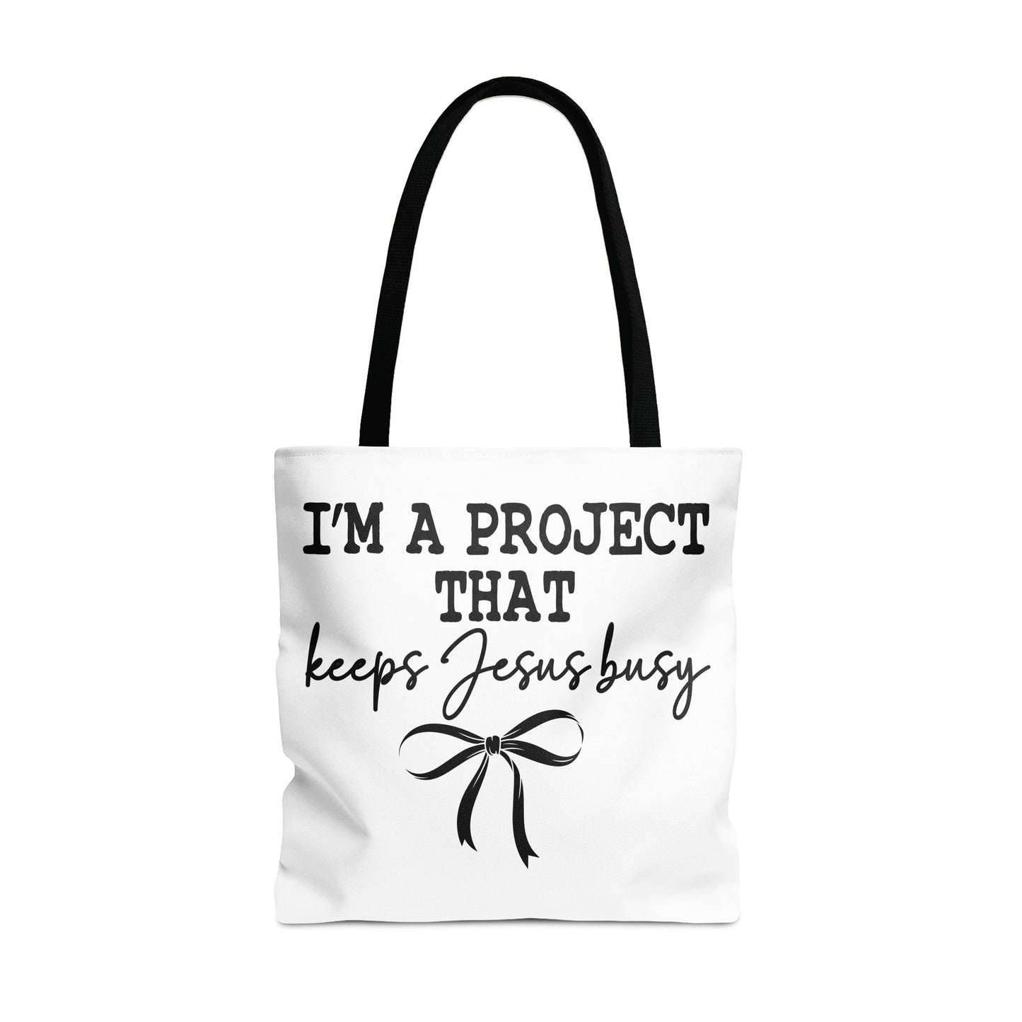 White tote bag with black text that reads "I'M A PROJECT THAT keeps Jesus busy" and a bow.