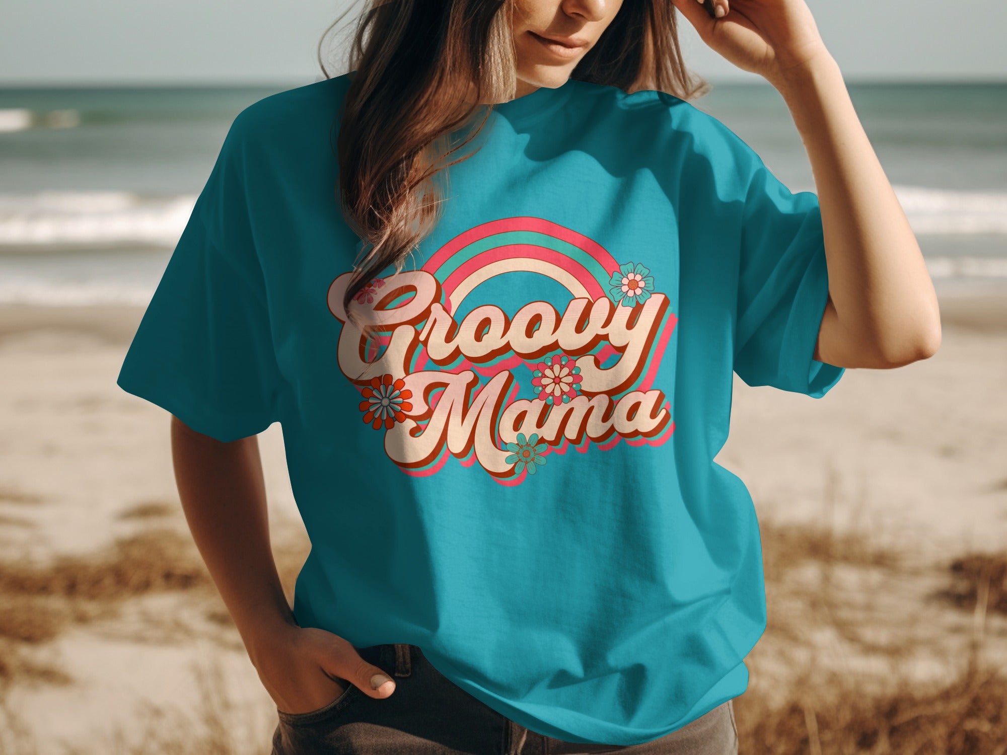 Teal t-shirt with "Groovy Mama" graphic and rainbow, set on a beach.