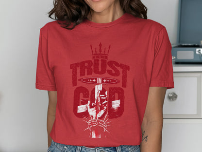 Red t-shirt with a graphic featuring a crown, text "TRUST IN GOD," and a hand holding a cross.
