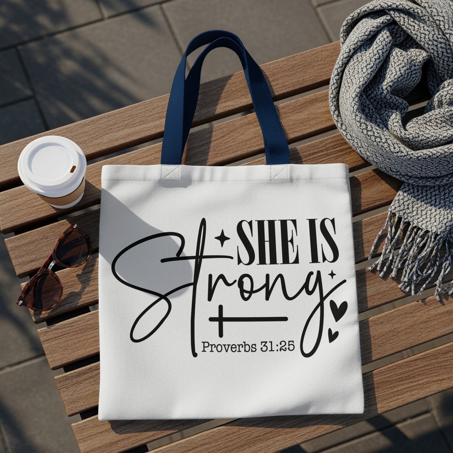 White tote bag with "SHE IS Strong + Proverbs 31:25" text on it.