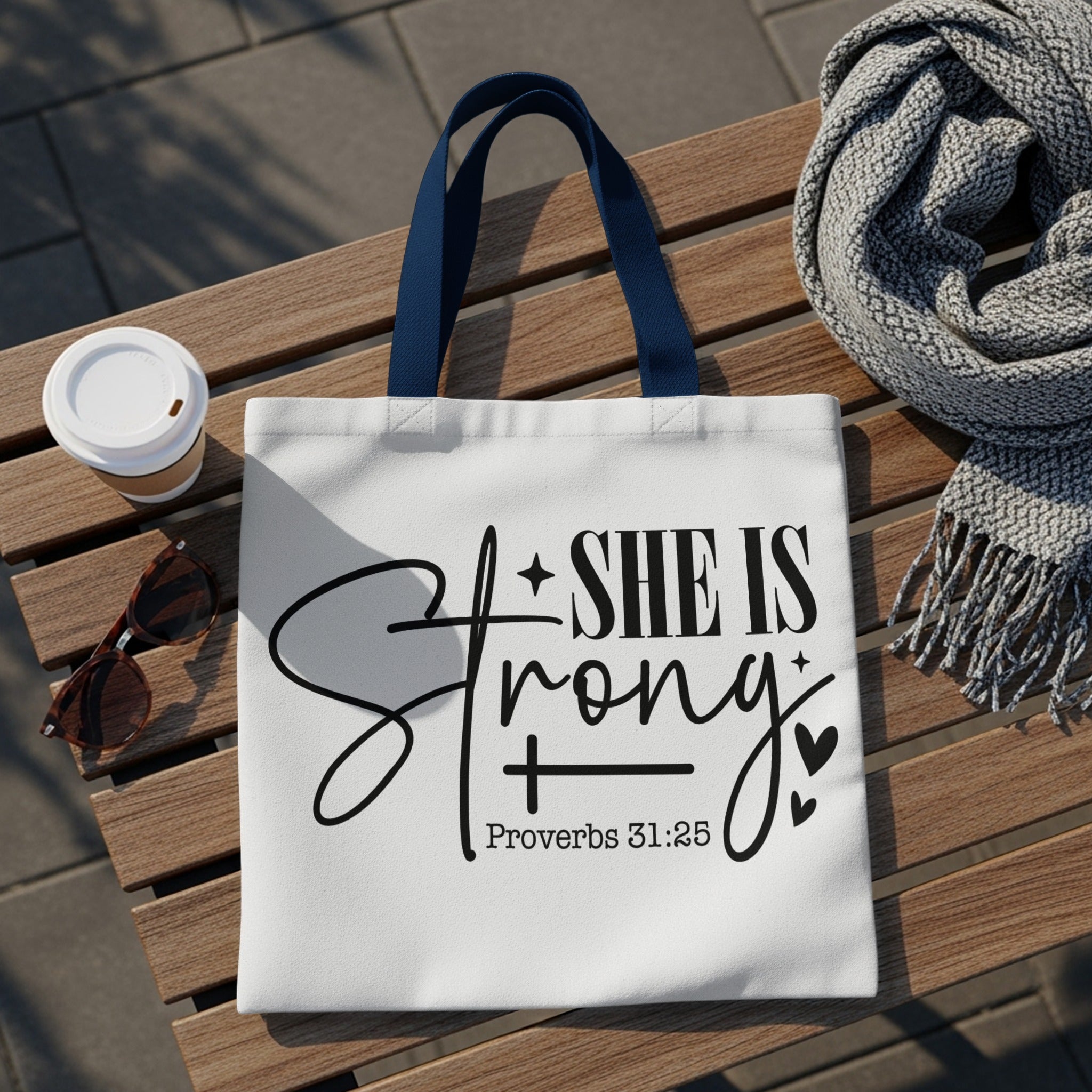 White tote bag with "SHE IS Strong + Proverbs 31:25" text on it.