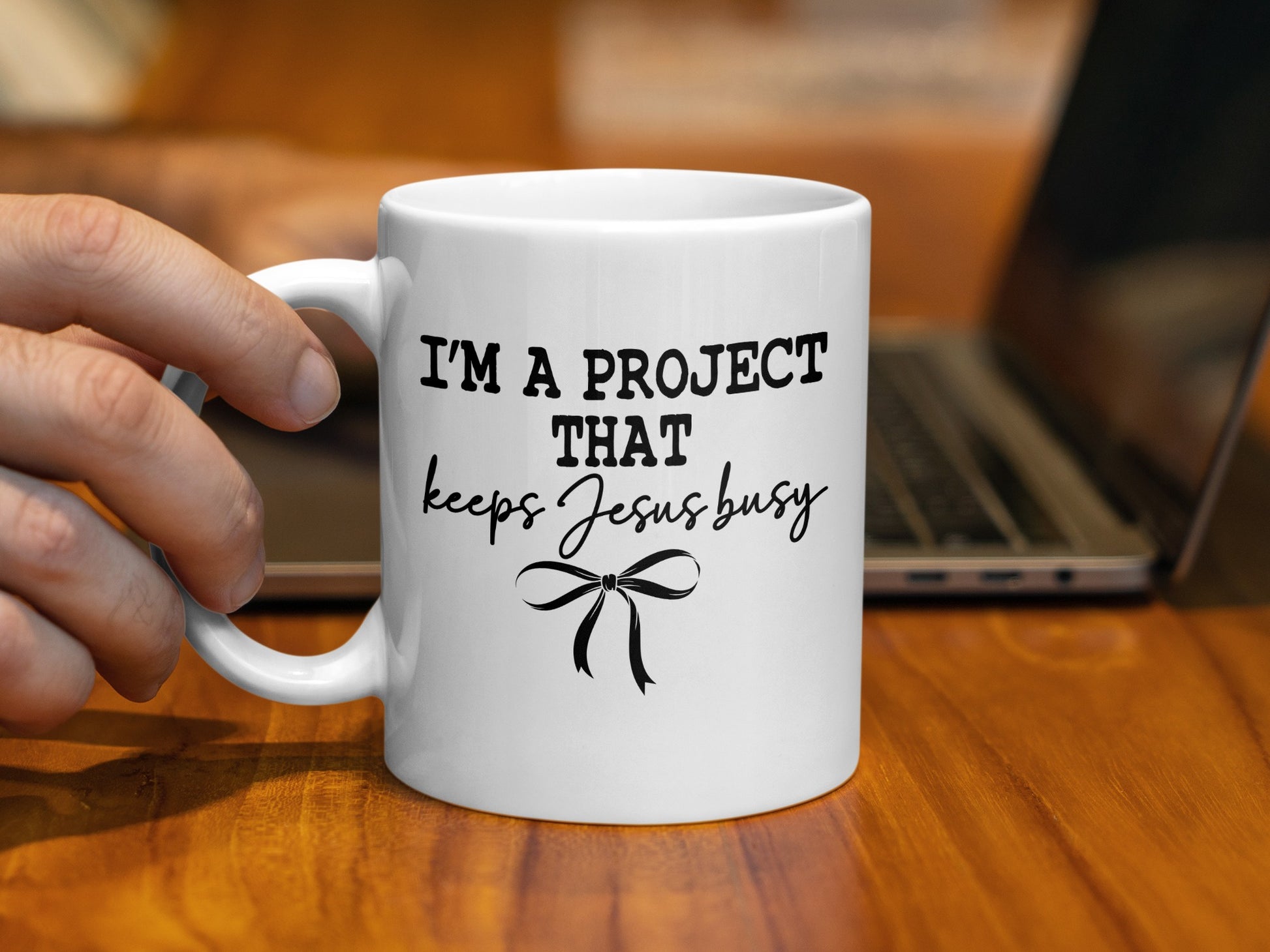 White mug with black text that reads "I'M A PROJECT THAT keeps Jesus busy" and a bow.
