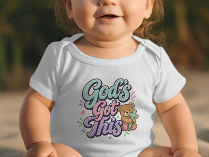 Baby wearing white bodysuit with text "God's Got This" and a teddy bear holding a cross.