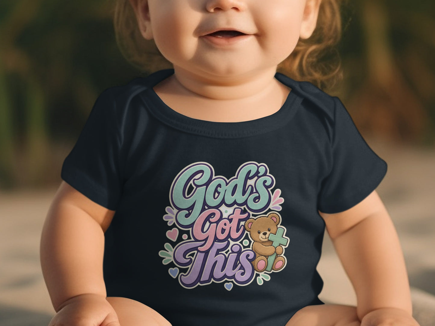 Baby in navy onesie with "God's Got This" text and teddy bear holding a cross.