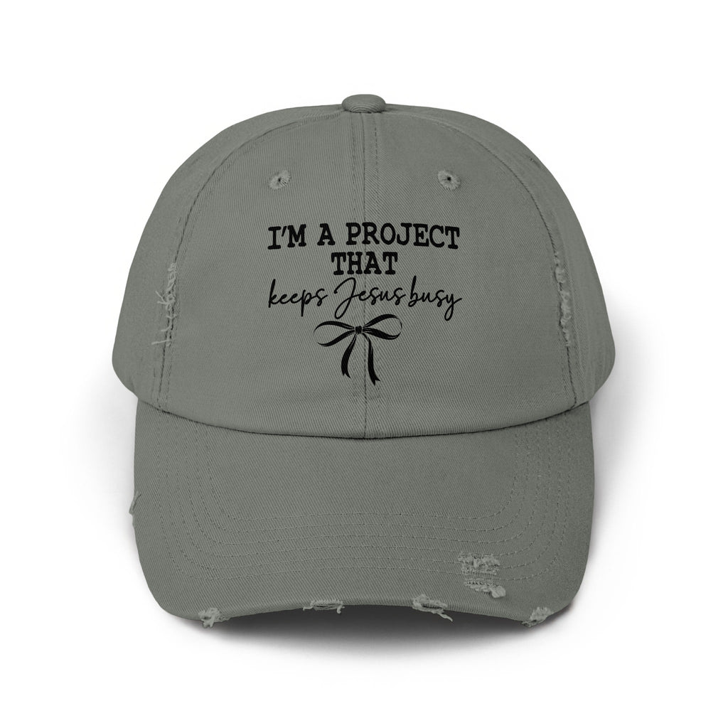 Distressed gray baseball cap with black text that reads "I'M A PROJECT THAT keeps Jesus busy" and a bow.