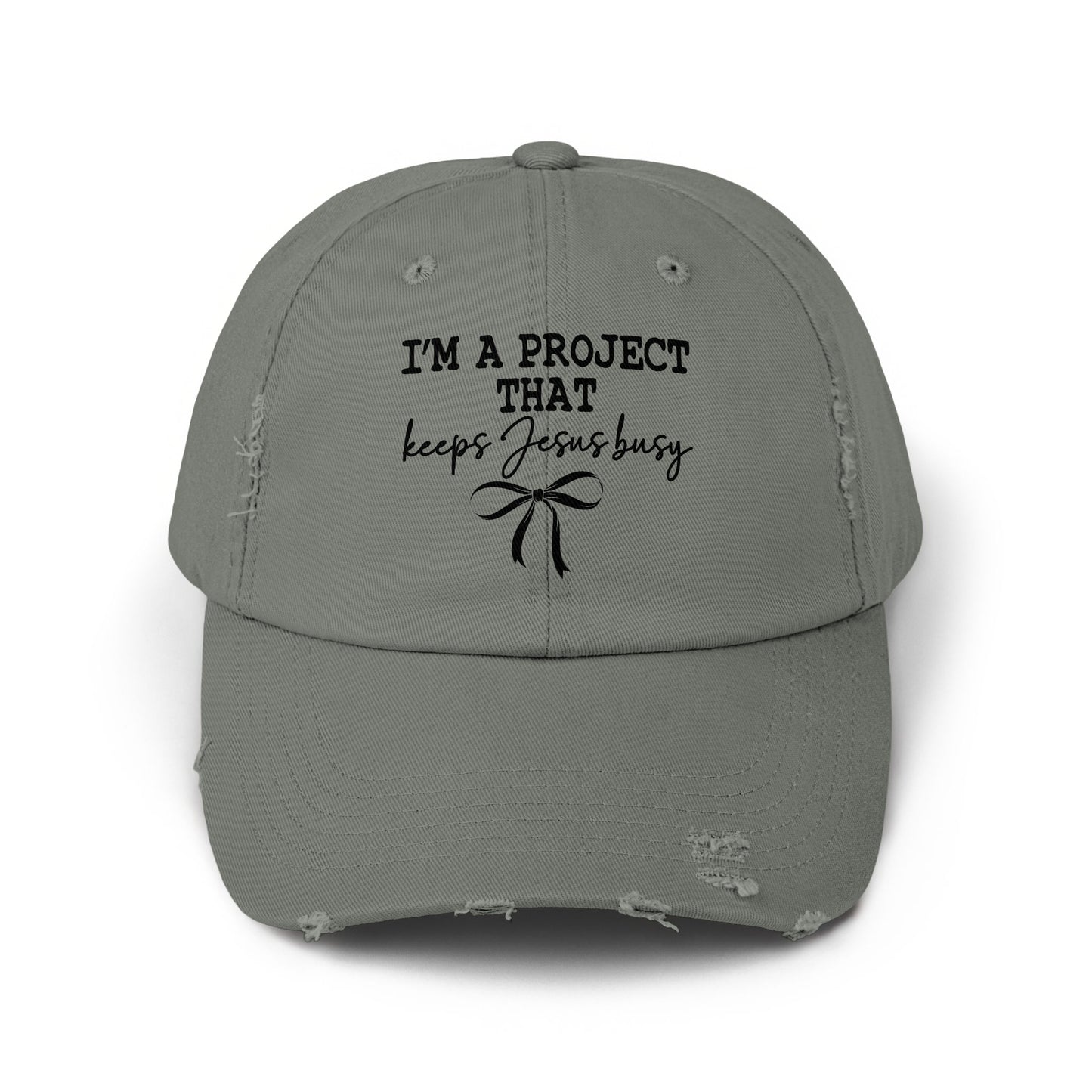 Distressed gray baseball cap with black text that reads "I'M A PROJECT THAT keeps Jesus busy" and a bow.