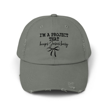 Distressed gray baseball cap with black text that reads "I'M A PROJECT THAT keeps Jesus busy" and a bow.