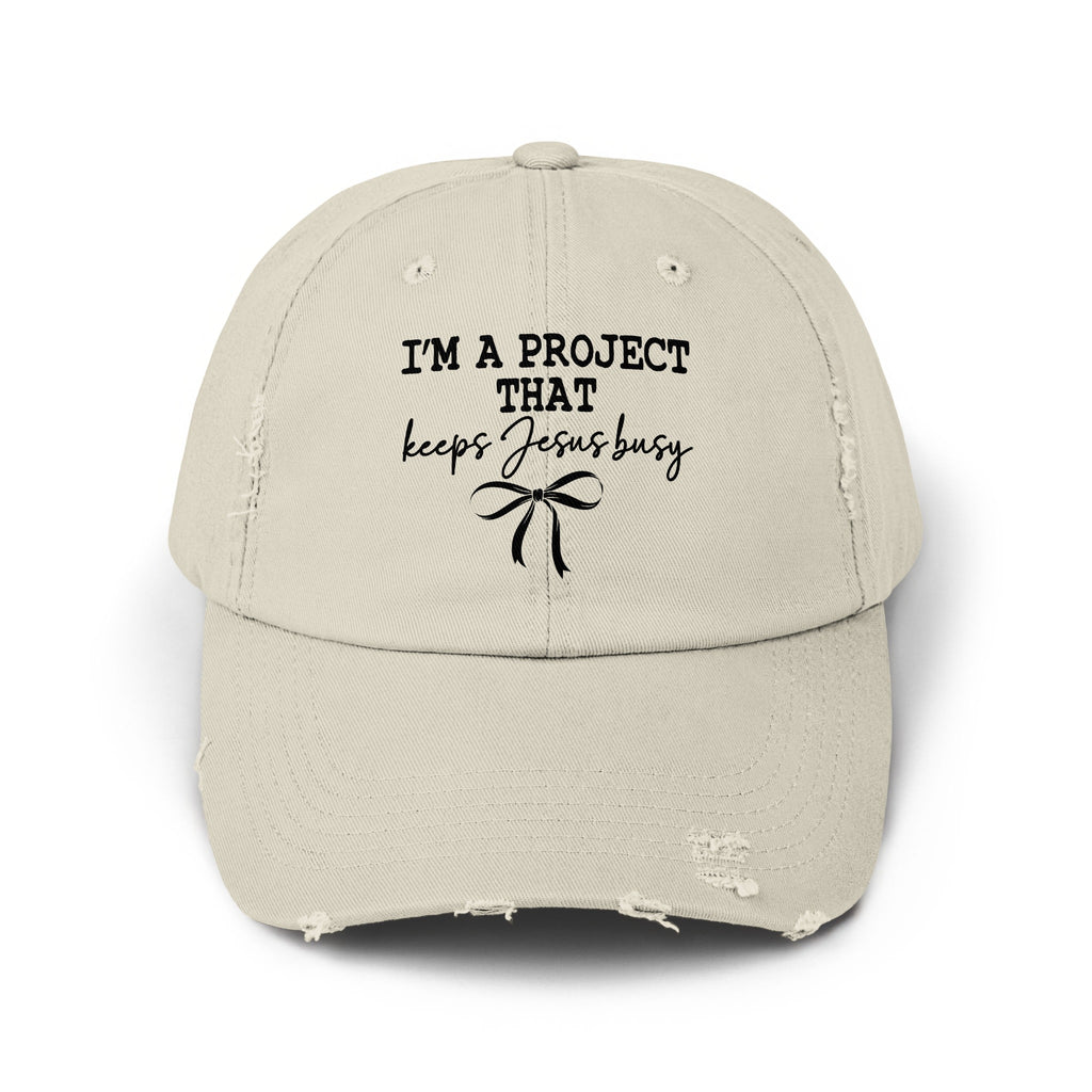 Distressed light beige baseball cap with black text "I'M A PROJECT THAT keeps Jesus busy" and bow.