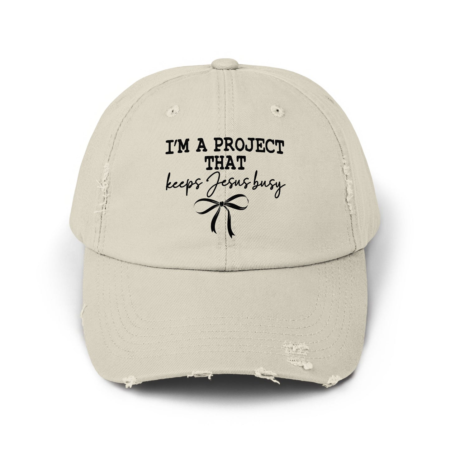 Distressed light beige baseball cap with black text "I'M A PROJECT THAT keeps Jesus busy" and bow.
