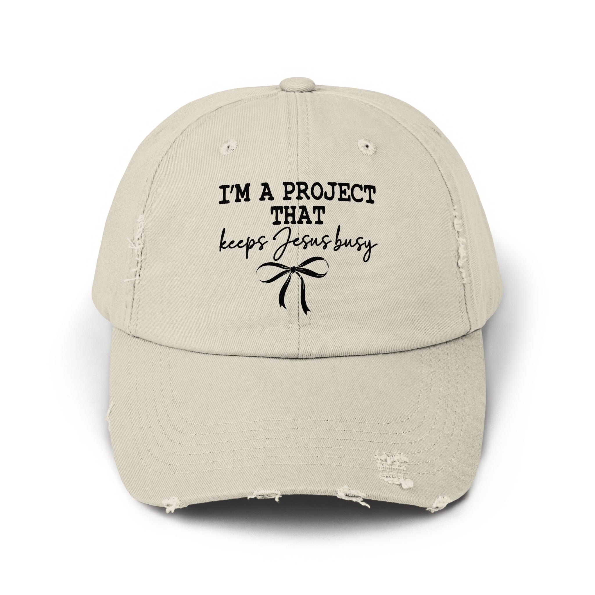 Distressed light beige baseball cap with black text "I'M A PROJECT THAT keeps Jesus busy" and bow.
