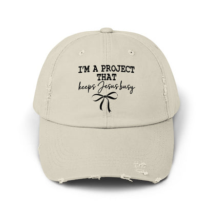 Distressed light beige baseball cap with black text "I'M A PROJECT THAT keeps Jesus busy" and bow.