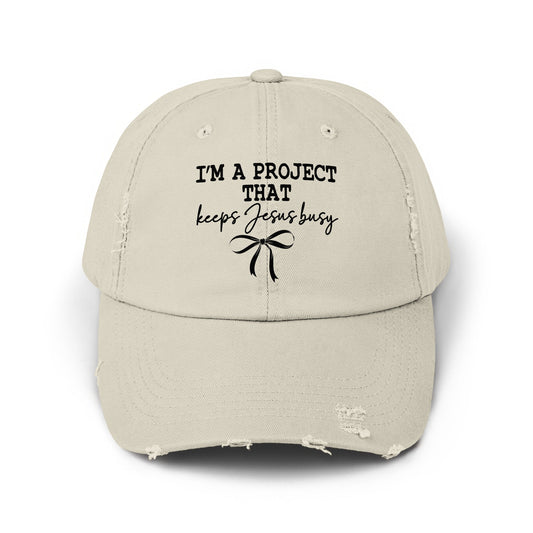 Distressed light beige baseball cap with black text "I'M A PROJECT THAT keeps Jesus busy" and bow.