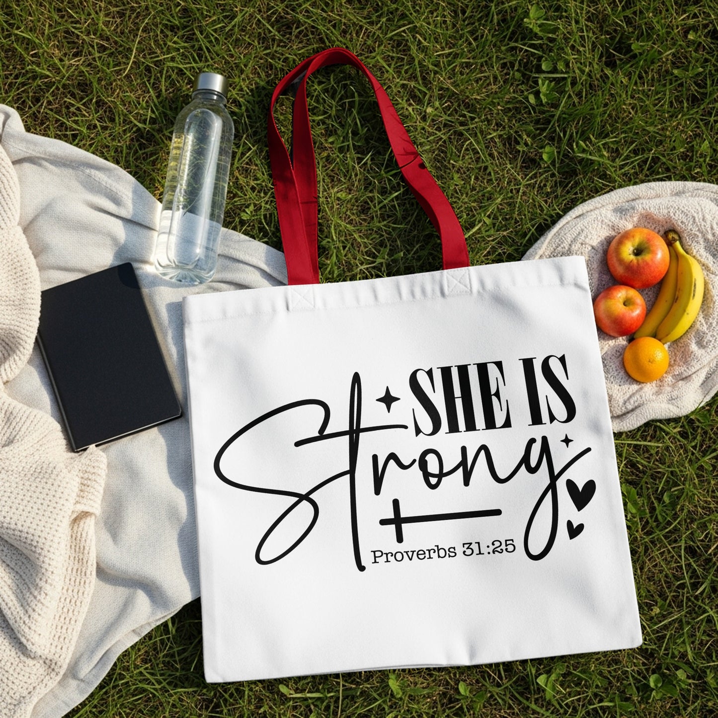 White tote bag with "She is Strong" and "Proverbs 31:25" text and fruit on grass.