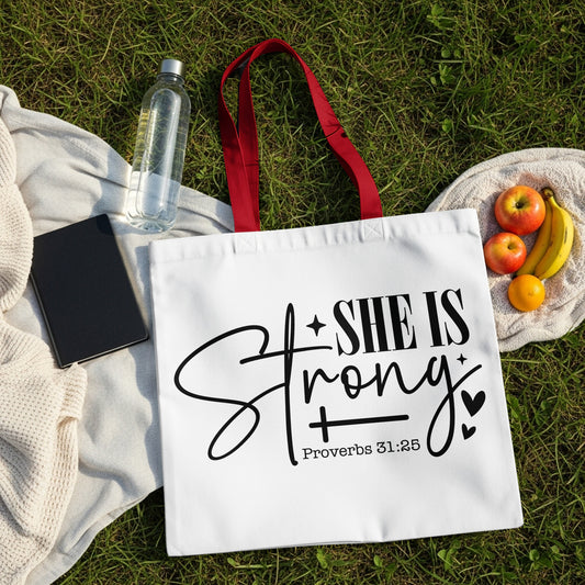 White tote bag with "She is Strong" and "Proverbs 31:25" text and fruit on grass.