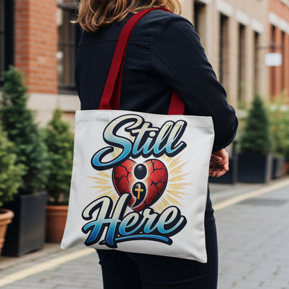 White tote bag with "Still Here" text and a cracked heart with a cross.