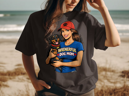 Woman in dark t-shirt holding dachshund, wearing red hat with paw print and blue shirt reading "WIENERFUL DOG MOM".
