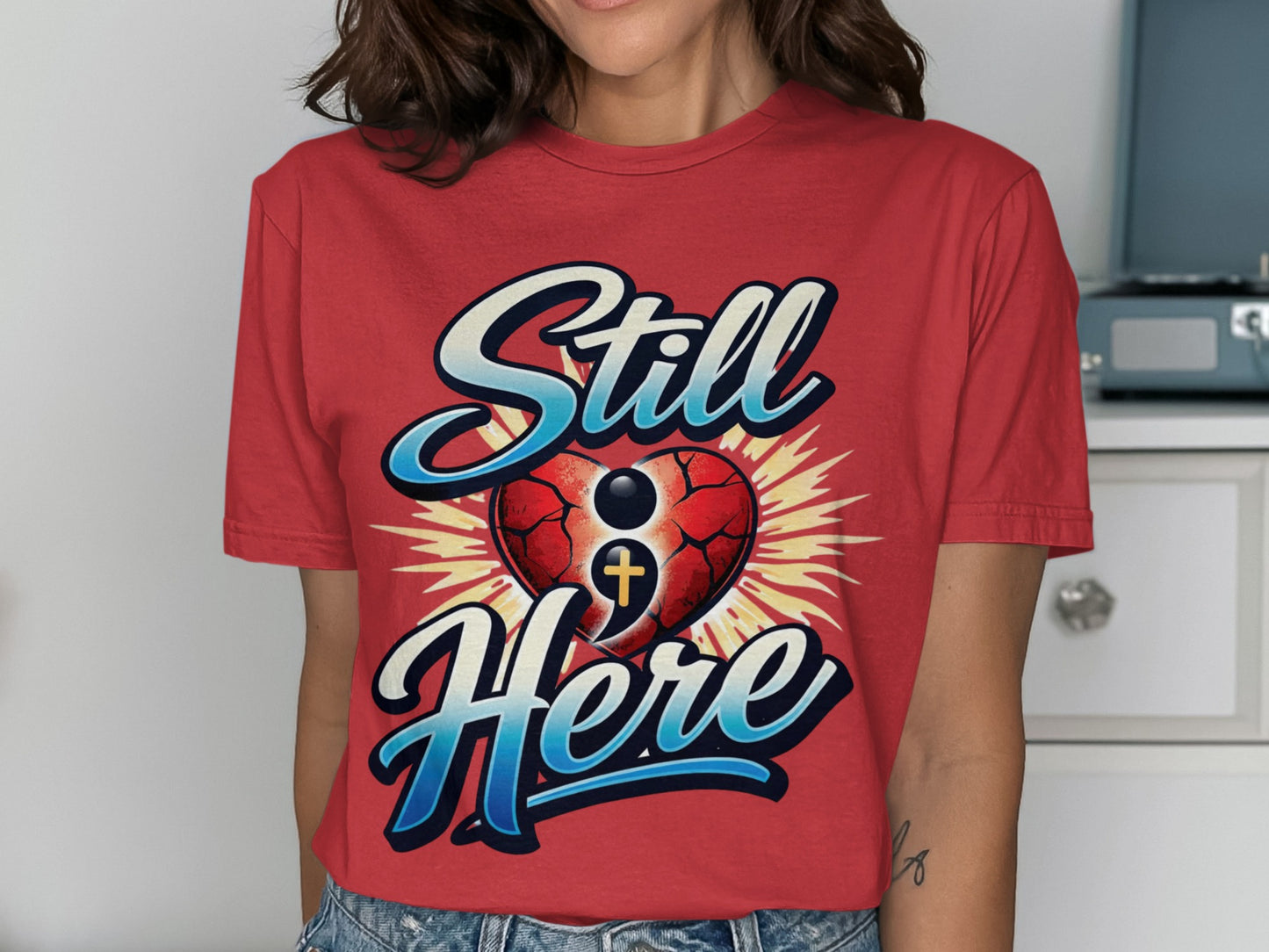 Red shirt with "Still Here" text, a cracked heart, and a semicolon cross symbol.