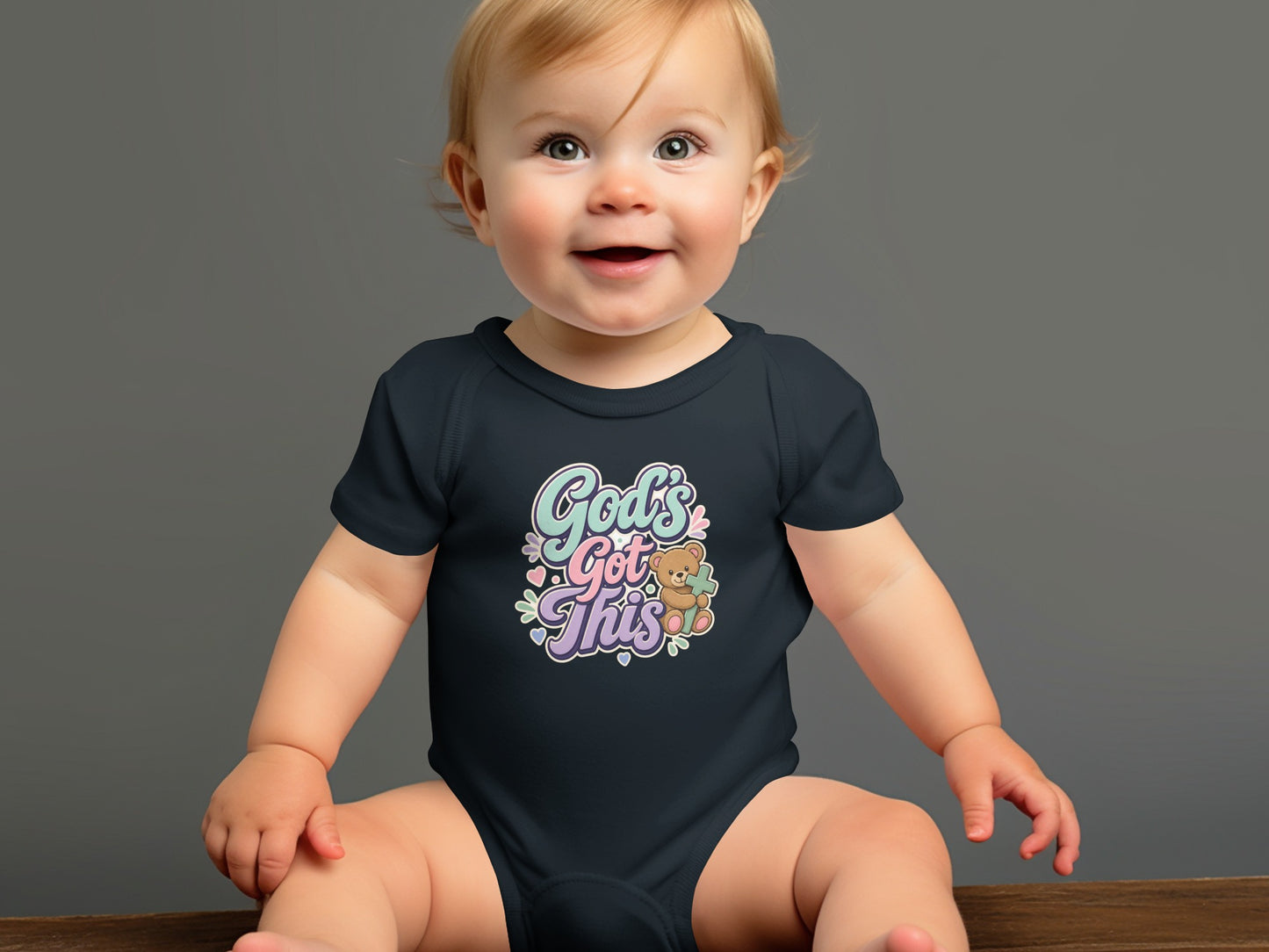 Baby wearing dark bodysuit with "God's Got This" text and teddy bear graphic.