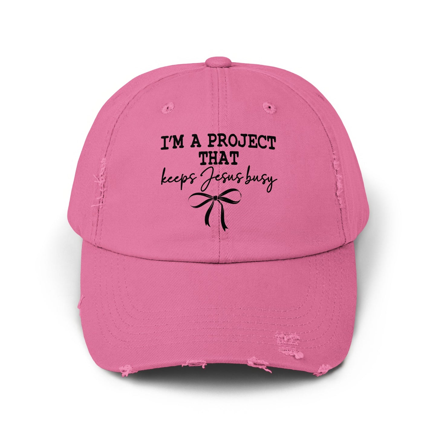 Pink distressed baseball cap with "I'M A PROJECT THAT keeps Jesus busy" text and bow.