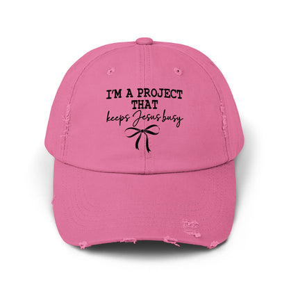 Pink distressed baseball cap with "I'M A PROJECT THAT keeps Jesus busy" text and bow.