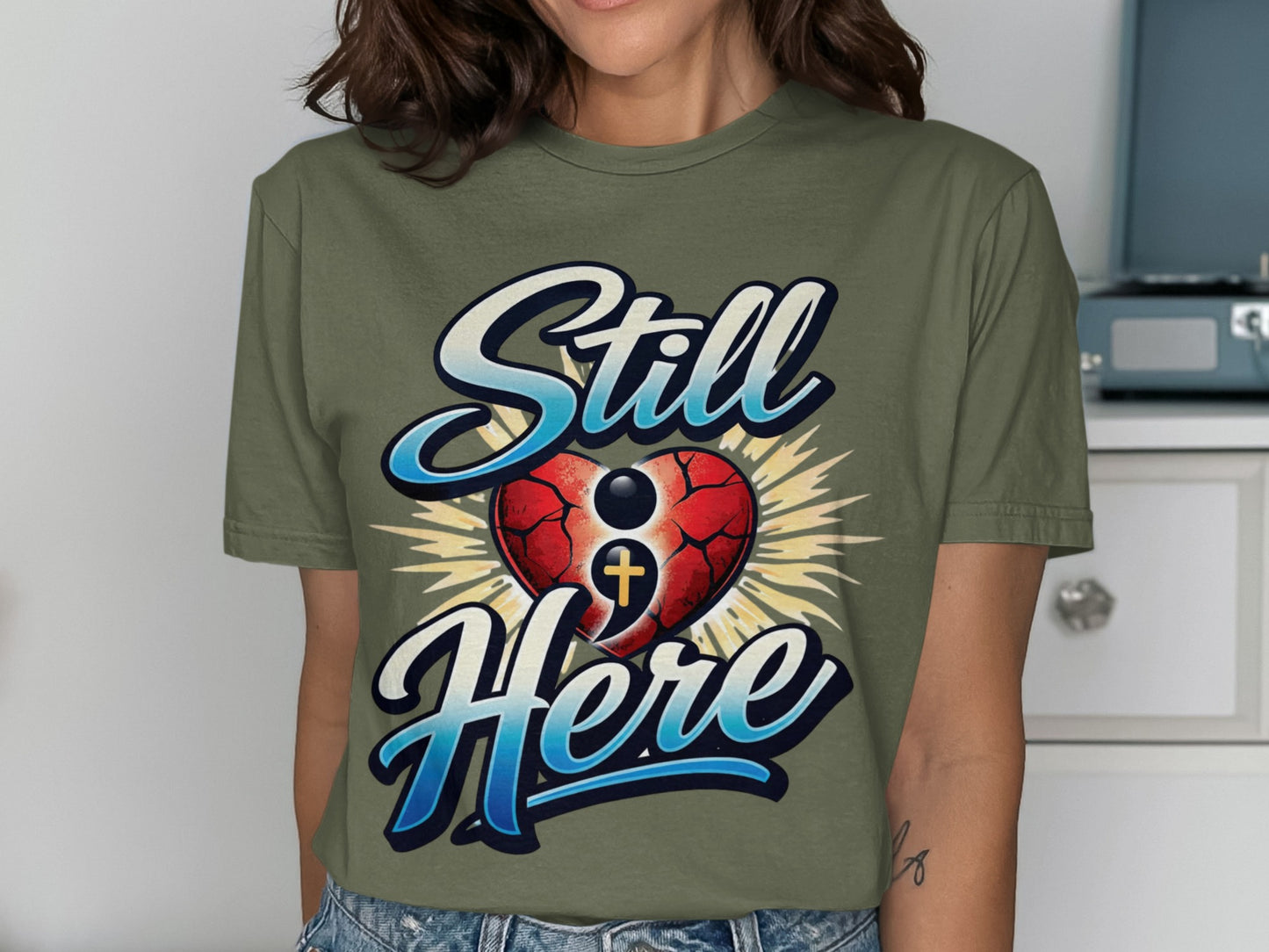 Green t-shirt with "Still Here" text and a symbolic heart with cross.