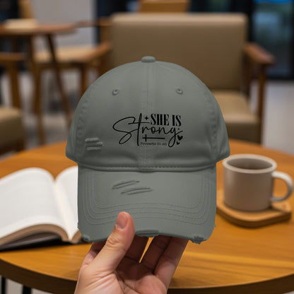 Gray distressed baseball cap with "SHE IS Strong" text and Proverbs 31:25.