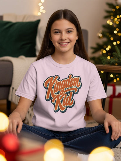 Girl in a pink t-shirt with "Kingdom Kid" text, seated with bokeh lights and Christmas tree.