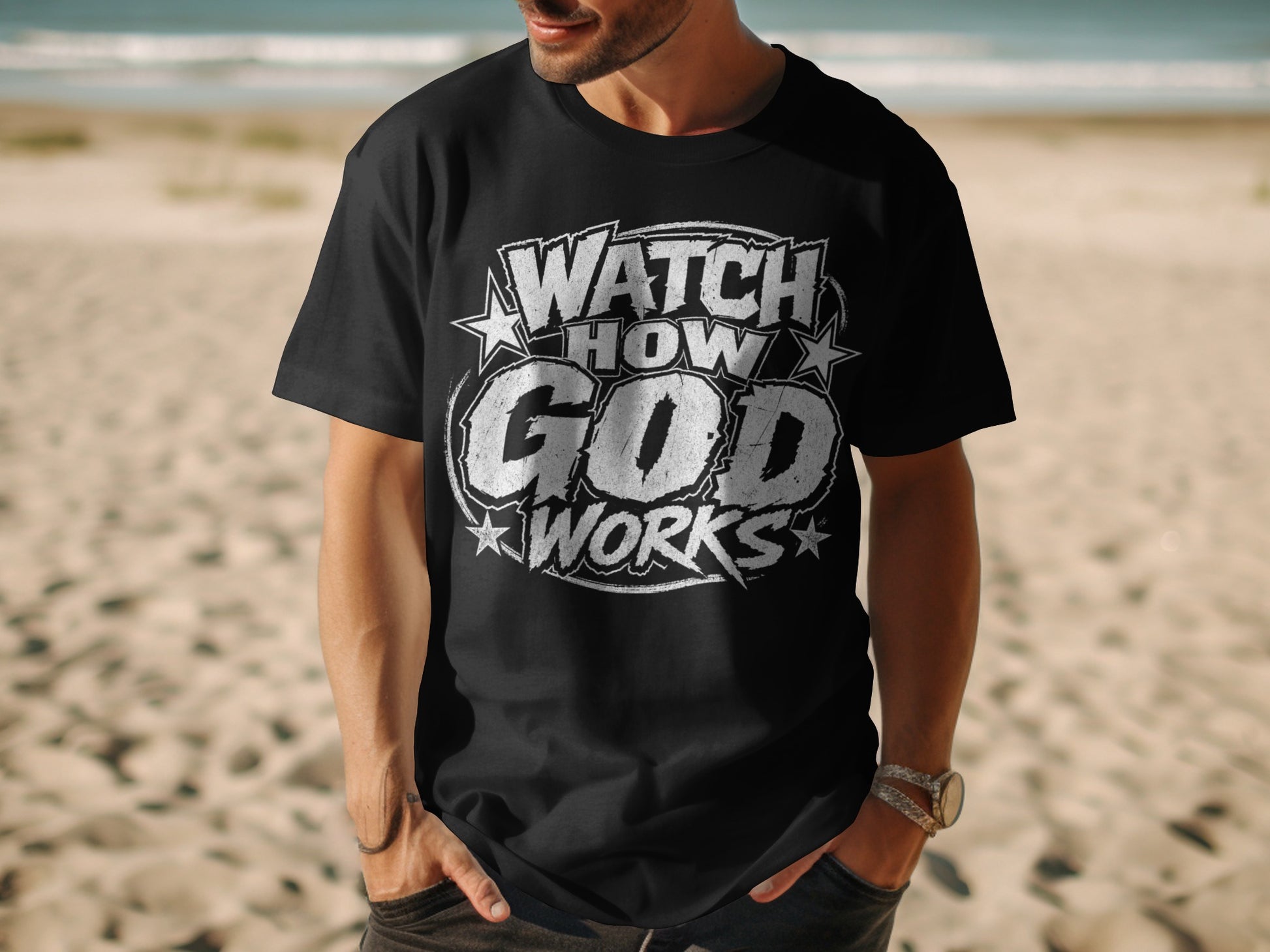 Black t-shirt with "WATCH HOW GOD WORKS" text design, worn by a person on a beach.