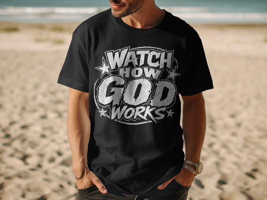 Black t-shirt with "WATCH HOW GOD WORKS" text design, worn by a person on a beach.