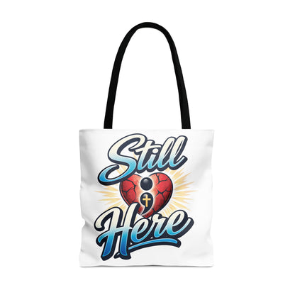White tote bag with "Still Here" text and a cracked heart with a semicolon and cross.